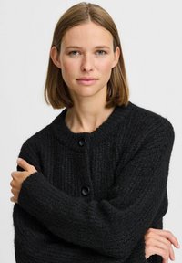 Čierny pletený cardigan s rebrovanou textúrou, okrúhlym výstrihom a tromi gombíkmi. Dlhé rukávy s uvoľneným strihom a širokými manžetami.