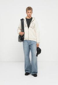 Joven vestido con una sudadera blanca de GAP con cremallera, camiseta negra, jeans de pierna ancha azules, zapatos negros, sosteniendo una bolsa negra y una gorra frente a un fondo blanco.