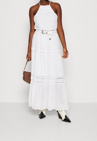 Bílá maxi šaty s halter výstřihem, krajkovým akcentem, stáhnuté v pase bílým opaskem. Nosené s černými botami na špičku a hnědou kabelkou.