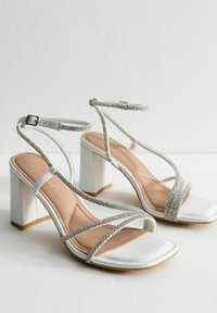 White high-heeled sandals with square toe, textured straps adorned with silver rhinestones, and a glossy block heel.