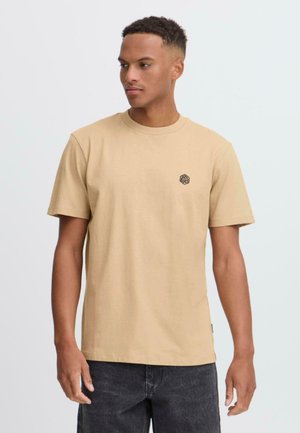 Beige bomulls T-skjorte med korte ermer, rund hals, med en liten svart geometrisk logo på venstre bryst. Myk tekstur, normal passform.