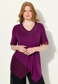Paarse tuniek met korte mouwen, V-hals en asymmetrische rand. Versierd met kleine studs bij de halslijn, gemaakt van zachte stof.