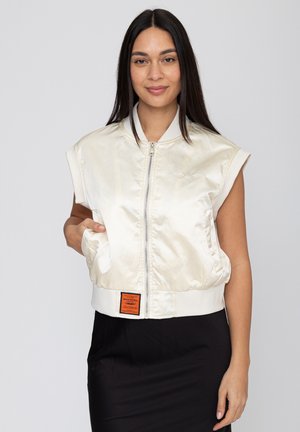 Vrouw die een mouwloze, glanzende witte vest met rits en zakken draagt, samen met een zwarte rok, staand tegen een effen lichte achtergrond.