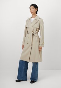 Beige trench coat with double-breasted buttons, large lapels, and a tie belt, worn over wide-leg blue denim jeans. Smooth texture and fitted design.