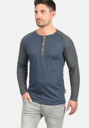 Majica s dugim rukavima henley stila s plavim tijelom i sivim raglan rukavima. Ima tri dugmeta na vratu i teksturiranu tkaninu.