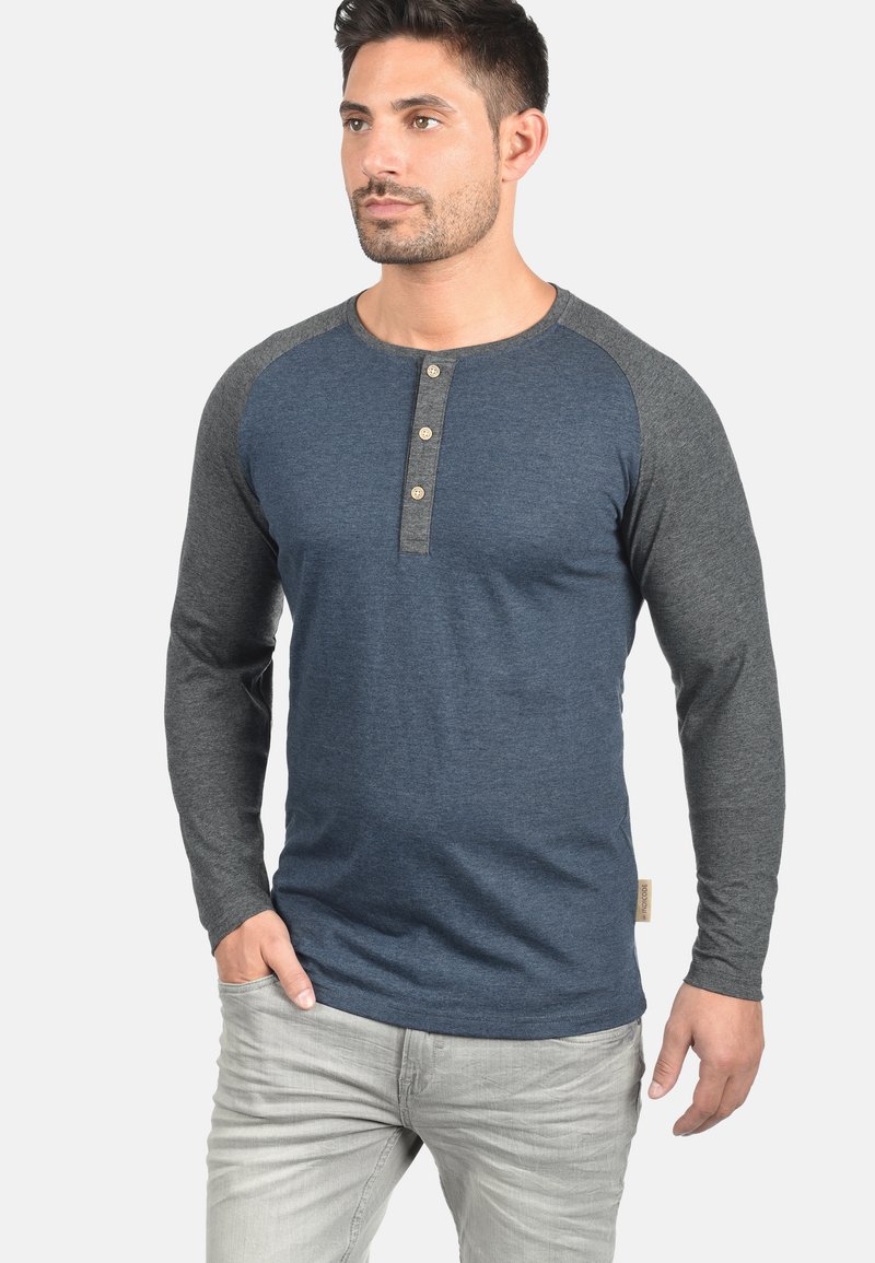 Langärmliges Henley-Shirt mit blauem Körper und grauen Raglanärmeln. Verfügt über drei Knöpfe am Hals und einen strukturierten Stoff.