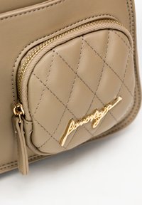 Borsa in pelle beige con texture trapuntata, dotata di zip dorata e un accentuato logo dorato. La cucitura dettagliata mette in risalto il design.