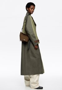Trench coat verde oliva con maniche larghe, dettagli marroni, abbinato a pantaloni beige e stivali neri. Inclusa una piccola borsa a spalla marrone.