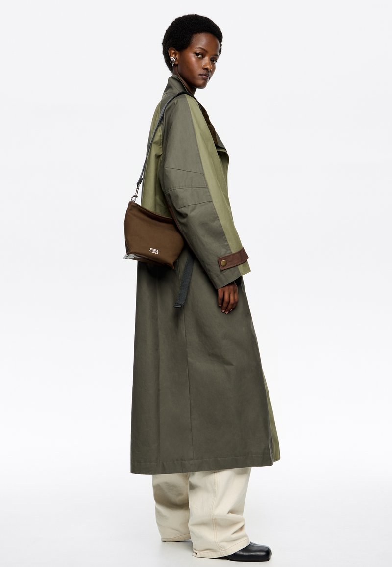 Trench coat verde oliva con maniche larghe, dettagli marroni, abbinato a pantaloni beige e stivali neri. Inclusa una piccola borsa a spalla marrone.