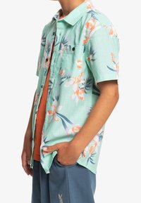 Kurzarmhemd aus mintgrünem Leinen mit floralen Mustern in Orange und Blau. Verfügt über eine Brusttasche und eine knöpfbare Blende.