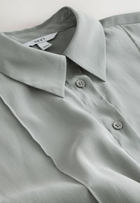 Jasnoszara koszula z kołnierzykiem i guzikami z przodu, z subtelnymi detalami szwów, ułożona na płasko, aby wyróżnić fakturę tkaniny i wzór.