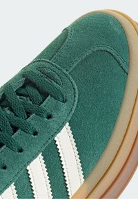 Zelené semišové tenisky s šněrováním, zdobené bílými cikcak pruhy a světle hnědou gumovou podrážkou.