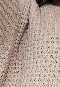 Närbild av en beige grovstickad tröja som bärs av en person, vilket lyfter fram det strukturerade, öppna stygnmönstret.