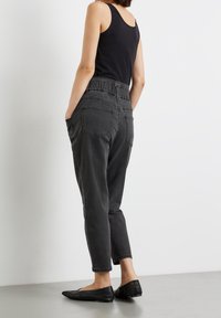 Mujer con top sin mangas negro, jeans oscuros de tiro alto con bolsillos traseros y zapatos planos negros de punta, de pie contra una pared blanca.