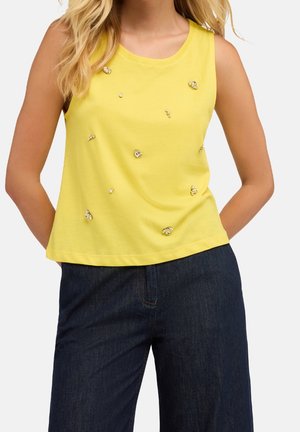 Haut jaune sans manches avec des petites ornements en cristal dispersées, porté avec un pantalon en jean bleu foncé.