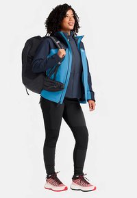 Blauw-zwart jack met ritssluiting, zwarte rugzak, strakke zwarte broek en rood-witte sneakers. De outfit heeft een sportief, gelaagd ontwerp.