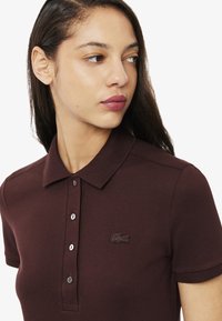 Polo shirt marron avec une texture, fermeture à trois boutons et un petit logo brodé sur la poitrine gauche. Manches courtes.