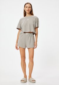 Grijze cropped t-shirt met korte mouwen en logo, gecombineerd met bijpassende shorts. Het model draagt zachte slippers. Eenvoudige textuur en een ontspannen pasvorm.