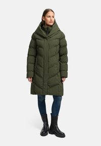 Olivegrøn pufferjakke med høj krave, prydet med diamantquiltning, lynlås og sidelommer, kombineret med blå jeans og sorte støvler.