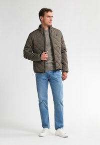 Gequilted olijfgroene jas met een hoge kraag, gecombineerd met een grijze trui en lichtblauwe jeans, compleet met witte sneakers.
