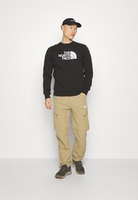 Sudadera negra de cuello redondo con logo blanco, pantalones cargo beige con cremalleras laterales y zapatos de senderismo grises, usados por una persona.