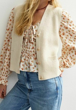 Femme portant un gilet en maille crème avec des boutons dorés sur un chemisier fleuri à manches longues et un jean bleu taille haute, sur un fond uni.