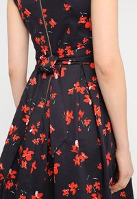 Ärmelloses dunkles Kleid mit rotem Blumenmuster und gebundenem Taillengürtel, von hinten gezeigt mit einem sichtbaren Arm.