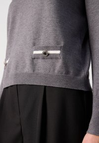 Grauer Strickpullover mit kontrastierenden weißen Streifen, vorderen Knöpfen und geripptem Saum, kombiniert mit schwarzen, plissierten Hosen.
