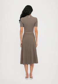 Femme aux longs cheveux noirs portant une robe marron moulante à manches courtes côtelée avec une jupe plissée, debout de dos sur un fond uni.