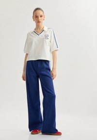 Witte polo met korte mouwen met marineblauwe strepen, v-hals en logo; gecombineerd met marineblauwe wijde broek en rode sneakers met bruine zolen.