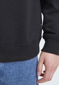 Schwarzer Sweatshirt mit gerippten Bündchen und Saum, mit einem seitlichen Etikettendetail, kombiniert mit hellblauen Denim-Jeans, die ein lässiges Outfit präsentieren.