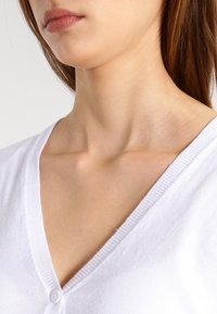 Bílý kardigan s hlubokým výstřihem ve tvaru V, texturovanou pleteninou, uzávěrem na jeden knoflík a žebrovanými okraji na krku a légě.