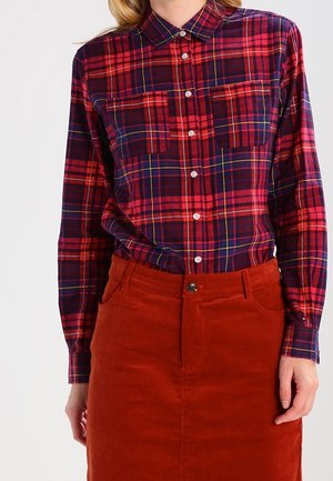 Kobieta ubrana w czerwoną i granatową koszulę w kratę, wpuszczoną w ciepłą rudą spódnicę ze sztruksu z przednimi kieszeniami.