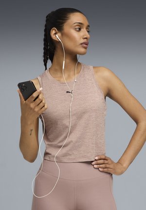 Haut court sans manches de couleur mauve clair avec un col rond et un logo Puma. Le modèle tient un smartphone noir et porte des écouteurs blancs.