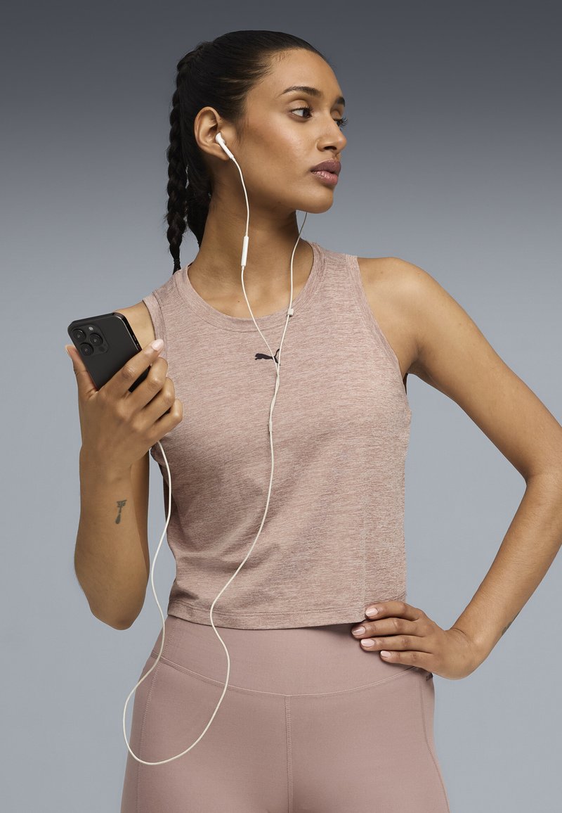 Φωτεινό μωβ αμάνικο crop top με στρογγυλή λαιμόκοψη και λογότυπο Puma. Το μοντέλο κρατάει ένα μαύρο smartphone και φοράει άσπρα ακουστικά.