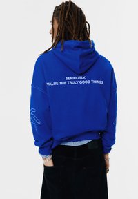 Blauwe hoodie met een grote witte opdruk aan de achterkant met de tekst "SERIEUS, WAARDEER DE ECHT GOEDE DINGEN" en witte handdesigneer omtrekken op de mouwen.