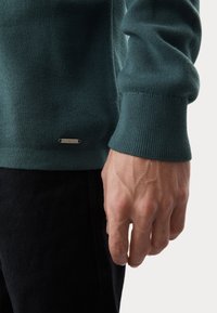 Turkusowy sweter z dzianiny z prążkowanymi mankietami, z małą metaliczną metką na dole, widoczny z ręką położoną na czarnym materiale.