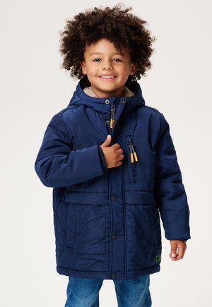 Lächelndes Kind mit lockigem Haar, das einen marineblauen Wintermantel mit Kapuze und Jeans trägt, vor einem einfarbigen Hintergrund stehend.