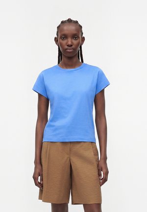 Femeie cu păr împletit, purtând un tricou bleu cu mânecă scurtă și pantaloni scurți maro largi până la genunchi, stând în fața unui fundal alb simplu.