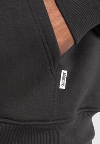 Sweat-shirt noir avec une texture côtelée, une poche kangourou, et une petite étiquette blanche marquée "SMILOOXX" sur la couture latérale.