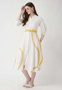 Weißes knielanges Hemdkleid aus strukturiertem Stoff, gelber Gürtel und geschwungene gelbe Akzente. Das Model trägt cremefarbene Sandalen und Blumenohrringe.