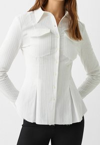 Woman wearing a fitted white button-up shirt with vertical stripes, flap chest pockets, and pleated sides, paired with black pants.