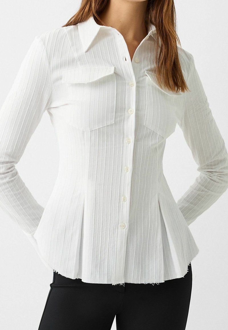 Woman wearing a fitted white button-up shirt with vertical stripes, flap chest pockets, and pleated sides, paired with black pants.