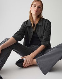 Zwarte textuur jacket, zwart topje en grijze flared jeans. Model zit gekruist, met zwarte instappers. Eenvoudige achtergrond.