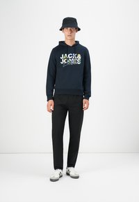 Marineblauer Kapuzenpullover mit mehrfarbigem "JACK & JONES"-Grafik, schwarze Hosen und ein schwarzer Bucket Hat. Weiße Schuhe mit sichtbaren grauen Akzenten.