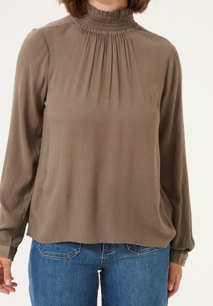 Kobieta w długim rękawie taupebluzki z marszczonym wysokim kołnierzem w połączeniu z niebieskimi dżinsami na jasnym tle.