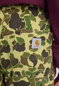 Geel camouflage cargo broeken met een achterzak, voorzien van een Carhartt logo label in oranje en wit, en een patroon in bruin, groen en zwart.