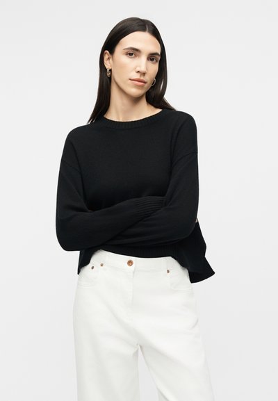 Γυναίκα με μακριά σκουρόχρωμα μαλλιά, που φοράει χρυσούς κρίκους σκουλαρίκια, μαύρο πουλόβερ με μακριά μανίκια και λευκό ψηλόμεσο παντελόνι με κουμπιά.