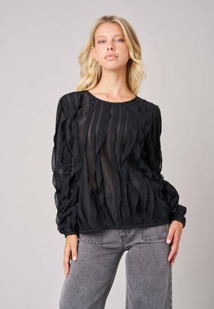 Blond vrouw die een doorschijnende zwarte blouse met lange mouwen draagt met verticale getextureerde strepen en grijze spijkerbroek tegen een effen achtergrond.