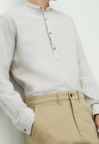 Camicia grigia chiara a maniche lunghe con colletto a band, pattina con bottoni e texture in lino. Abbinata a pantaloni beige con tasche laterali.